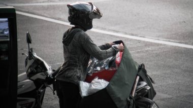 Kask takan bir teslimatçı, motosikletlerine büyük yeşil bir kargo çantası yüklüyor. Sahne sessiz, monokromatik bir şekilde çekilir, şehir ortamını ve sürücünün görevini vurgular..
