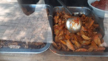 İki metal tepside Endonezya yemekleri sergileniyor: Tempeh ve baharatlı patates ve kepçe. Yiyecekler camın arkasında, tahta bir yüzeyde..