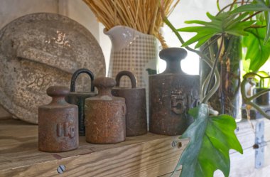 Kırsal bir mutfak rafında klasik metal ağırlıklar, buğday saplı bir seramik sürahi ve yapraklı bir bitki bulunur. Ağırlıklar kilogramlarla işaretlenir ve sahneye nostaljik bir dokunuş eklenir..