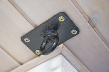 Tahta bir yüzeye monte edilmiş, sarı vidalarla korunmuş siyah metal bir tavan kancasının yakın çekim görüntüsü. Kanca, eşyaları güvenli bir şekilde asmak için tasarlandı..