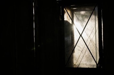 Çaprazlama desenli loş bir pencere. Gölgelerle kısmen gizlenmiş. Az miktarda ışığın geçmesine olanak sağlıyor..