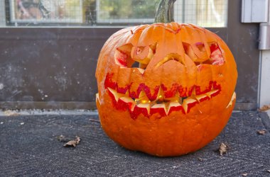 Korkunç yüzlü oyulmuş bir balkabağı, sivri dişli ve içinde parıldayan ışıklar olan, bir kapı eşiğine yerleştirilmiş.