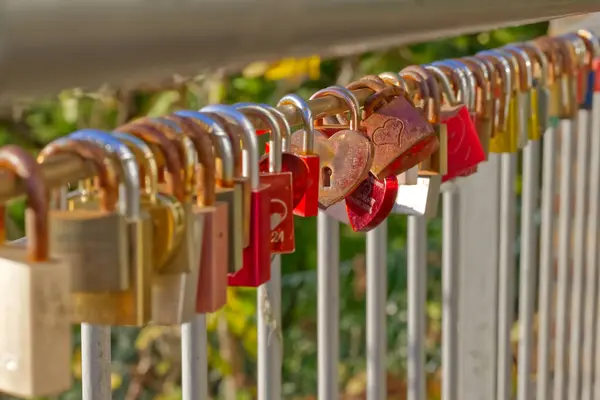 Metal korkuluklara bağlanmış renkli asma kilitler, aşkı ve bağlılığı sembolize ediyor. Arka planda tuğla bir duvar ve bulanık yeşillik var. Romantik bir atmosfer yaratıyor..