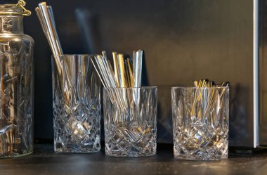 Karanlık bir yüzeye yerleştirilmiş metal pipetlerle dolu kristal bardak koleksiyonu. Gözlükler farklı boyutlarda, karmaşık tasarımlar sergiliyor ve ışığı güzel yansıtıyor..
