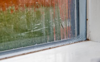 Yağmura ıslanmış bir pencerenin yakından çekilmiş görüntüsü, su damlacıklarını ve camdaki çizikleri gösteriyor. Bulanık arka plan yeşilliği yansıtıyor, sakin bir atmosfer yaratıyor..