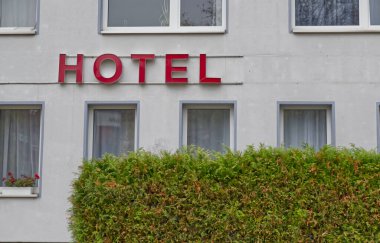 Kırmızı 'HOTEL' işareti olan bir otel binası. Ön cephe, beyaz perdeli çok sayıda pencereye sahip açık renkli. Önünde yeşil bir çit görünür, biraz doğa katar..
