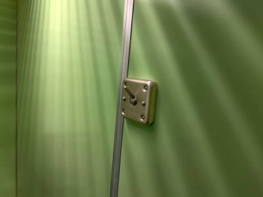 Yeşil bir duvarda metalik bir mandalın yakın çekim görüntüsü modern bir tasarımı parlak bir kaplamayla sergiliyor. Arka planda, yüzeyde gradyan etkisi yaratan yumuşak ışıklandırma var.