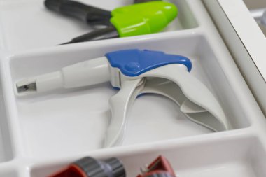 Alet düzenleyicisinin kompartımanına yerleştirilmiş beyaz ve mavi bir tetiğe yakın çekim görüntüsü. Arka plandaki diğer araçlar kısmen görülebilir olup, buna yeşil saplı ve çeşitli eklentiler de dahildir.