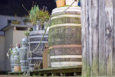 Ahşap fıçıların olduğu kırsal bir sahne, bazıları yosun kaplı, açık havada düzenlenmiş. Saksı bitkileri varillerin üstüne yerleştiriliyor ve biraz yeşillik katıyor. Dekorun doğal, dünyevi bir havası var. Ön planda tahta bir direk var..