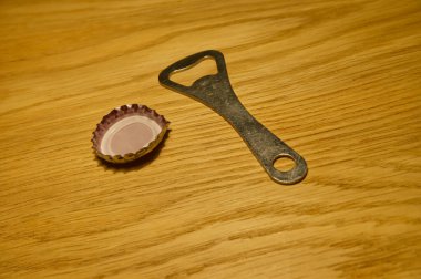 Ahşap bir yüzeydeki şişe kapağının yanında metal bir şişe açacağı. Açacak, yuvarlak uçlu klasik bir tasarıma sahipken, kapak hafif buruşuk ve mor kenarlı.