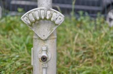 Yıpranmış metal bir direğin yakın çekim görüntüsü, uzun otlarla çevrili kavisli bir gösterge. Kutup daha büyük bir yapının parçası gibi görünüyor, muhtemelen yangın musluğu ya da benzer bir ekipman..