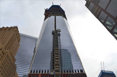 İnşaat halindeki uzun bir gökdelen, cam bir cephe ve iskele sergiliyor. Bina, üzerinde bulutlu bir gökyüzü bulunan diğer modern binalarla çevrilidir..