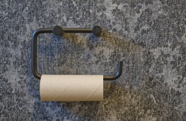 Minimalist siyah bir tuvalet kağıdı tutacağı, içi boş bir karton rulo ile dokulu gri bir duvara monte edilmiş..