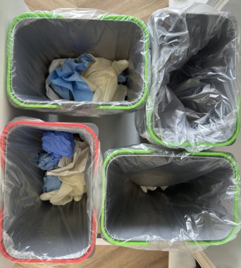 Renkli kenarlı dört çöp kutusu, her biri kullanılmış eldiven içeriyor. Çöp kutuları temiz plastik poşetlerle kaplı ve şebeke şeklinde düzenlenmiş..
