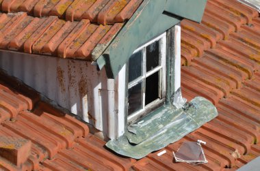 Küçük bir yurt penceresi olan eski, yıpranmış bir çatı. Pencere boyası soyulmuş ve kısmen açık, karanlık bir iç mekan ortaya çıkıyor. Çevredeki fayanslar kırmızı ve aşınma izleri var, biraz da yosun var..