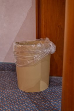 Plastik kapaklı bej bir çöp tenekesi, ahşap bir kapının yanında desenli bir halıda..