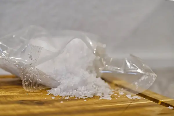 Temiz bir plastik torba ahşap bir yüzeye kaba beyaz tuz dökerek dokulu bir kontrastı yaratır. Arka plan hafifçe bulanık, tuz ve poşeti vurguluyor..