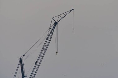 Gri gökyüzüne karşı asılı uzun bir inşaat vinci. Vinç yapısı siluetlidir, boyunu ve endüstriyel tasarımını vurgular..