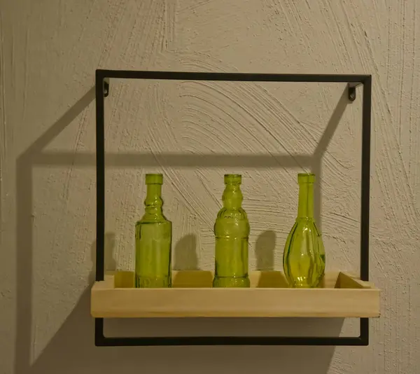 Tahta bir zeminde üç yeşil cam şişe bulunan minimalist bir duvar rafı. Rafta siyah metal bir çerçeve var, desenli gri bir duvara dayalı..