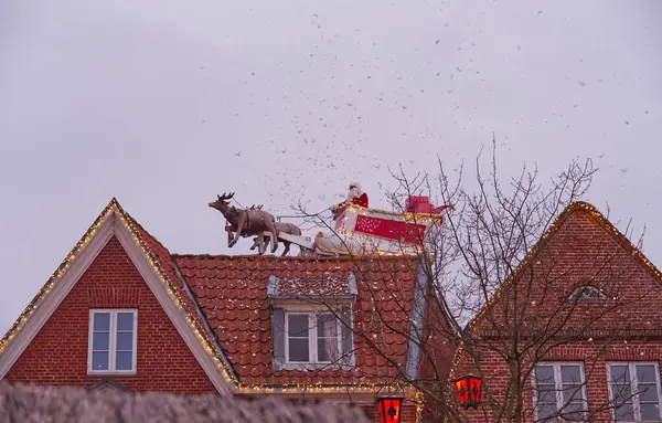 Noel Baba 'nın çatıda ren geyiğine bindiği ve kar tanelerinin yağdığı tuhaf bir sahne. Evler şenlikli ışıklarla süslenmiş, neşeli bir tatil ortamı yaratıyor..