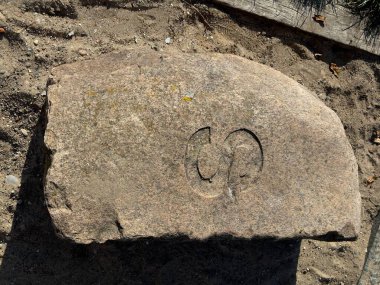 Büyük, düz bir taş, oyulmuş bir sembol ile stilize bir harf 'U' ya veya benzer bir şekle benziyor. Taş kumlu zeminde duruyor, doğal elementlerle çevrili..