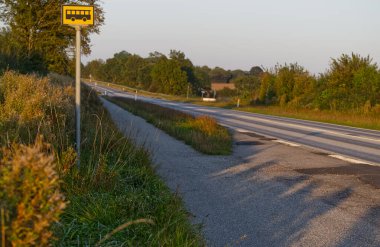 Etrafı çimen ve ağaçlarla çevrili otobüs durağı işareti olan kırsal bir yol. Güneş batıyor, kaldırımda uzun gölgeler bırakıyor. Manzara huzurlu ve sessiz, kırsal alanların tipik bir örneği..