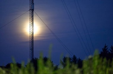 Yüksek bir iletişim kulesi gece parlak bir dolunaya karşı siluetlendi. Arka planda elektrik hatları ve ön planda çimenler vardı..