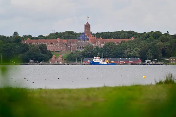 Marinefernmeldeschule 'un Flensburg' daki Strategische Aufklaerung için bir tepenin doğu kıyısındaki ağaçlarla çevrili, yakınlarda bir teknesi olan sakin bir su gövdesine bakan manzarası. Gökyüzü bulutlu..