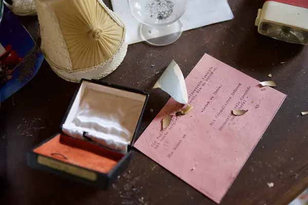 Kırık bir porselen parçası, bir mücevher kutusu ve pembe bir belgeyle terk edilmiş bir masa. Sahne, toz ve dağınıklıkla nostalji ve ihmalkarlık hissi veriyor..