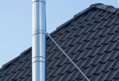 Koyu renkli bir çatıya bağlı parlak metal bir baca borusunun yakın çekim görüntüsü, arka planda açık mavi bir gökyüzü var..