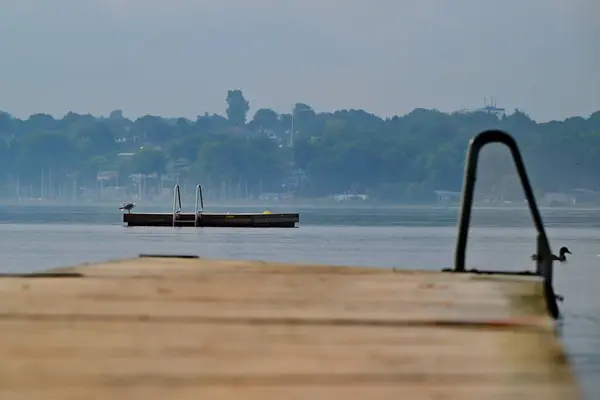 Kollund Danimarka 'da sakin sulara uzanan, uzakta küçük bir platforma sahip ahşap bir rıhtım manzarası. Arka planda ağaçlar ve sisli bir ufuk çizgisi var, sakin bir atmosfer yaratıyor..