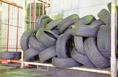 Depoda metal bir rafa yığılmış büyük bir kullanılmış lastik yığını. Lastikler çeşitli boyutlarda ve yıpranma izleri var, bazıları rastgele dizilmiş..