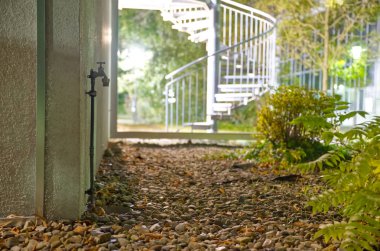 Arka planda sarmal bir merdiven bulunan sakin bir açık hava sahnesi, yemyeşil ve kayalık bir patikayla çevrili. Ayar hafifçe aydınlatılıyor, sakin bir atmosfer yaratıyor..