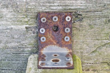 Yıpranmış ahşap bir yüzeye bağlı paslı metal bir parantez yakın plan, birden fazla vida ve merkezde bir delik var..