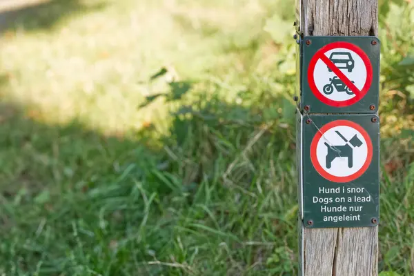 Park ya da açık alan kurallarını gösteren üç işareti olan ahşap bir direk. Tabelalar araçları ve motosikletleri yasaklıyor ve köpeklerin bir ipucunun üzerinde olmasını gerektiriyor. Metin, Danca ve Almanca da dahil olmak üzere birden fazla dilde yazılmıştır..