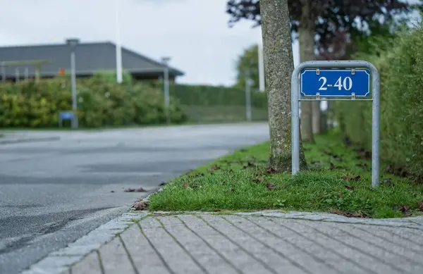 Ağaçlar ve yeşilliklerle kaplı sessiz bir yolun arka planına karşı 2-40 rakamlarını gösteren bir sokak tabelası. Sahne huzurlu bir banliyö atmosferini yakalıyor..