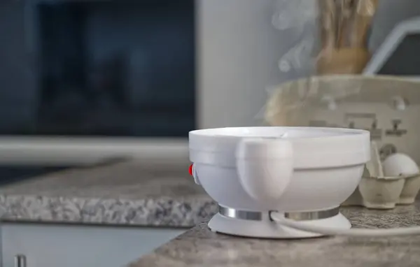 Mutfak ortamında granit bir tezgaha yerleştirilmiş, buhar yayan beyaz bir elektrikli yumurta fırını. Arka planda yumuşak ışıklandırmalı bulanık bir mutfak ortamı var..
