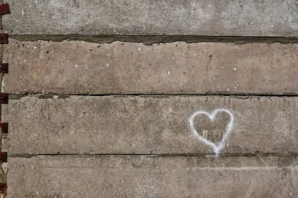 Tebeşirle çizilmiş basit bir beyaz kalp şeklindeki desenli beton bir duvar. Duvarda gri ve bej renklerin karışımı var. Görünür kusurları ve paslanmış metal elementleri var..