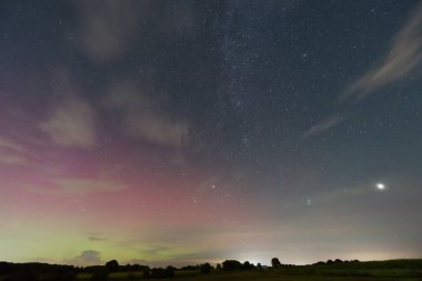 19 Eylül 2023 'ten kalma yıldızlarla dolu göz kamaştırıcı bir gece gökyüzü. Yeşil ve pembenin tonlarında bir aurora borealis. İnce bulutlar sahnede sürükleniyor, sağ tarafta parlak bir gezegen görünüyor..