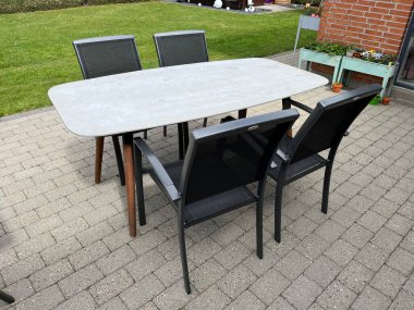 Dikdörtgen gri masa üstü ve dört siyah sandalyesi olan modern bir açık hava yemek masası. Masa çimlerle çevrili asfalt bir veranda ve arka planda tuğla bir duvar üzerine kurulmuş..