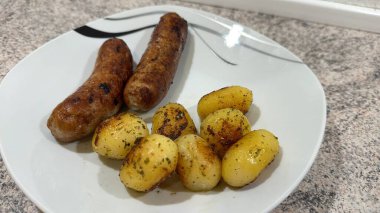 İki ızgara sosis ve bir porsiyon altın sarısı fırında patates, otlarla süslenmiş..