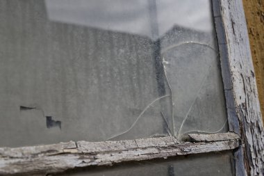 Kırık camlı ve soyulmuş boyalı yıpranmış bir pencerenin yakın çekimi. Toz ve kir camda görünüyor, bulanık bir arkaplanı yansıtıyor..
