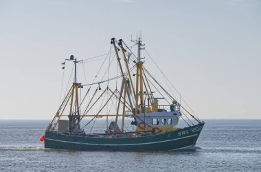 Sakin sularda yüzen bir balıkçı teknesi, yeşil gövdesini ve balıkçı malzemelerini sergiliyor. Tekne çeşitli anten ve bayraklarla donatılmıştır, açık mavi gökyüzüne karşı dizilmiştir..