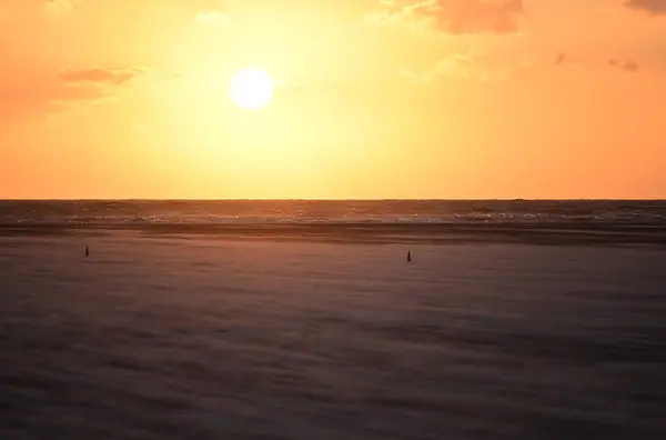 Güneş kuzey denizi üzerinde batarken, kumsalda bir kum fırtınası ufukta güneş batarken ve sıcak turuncu ve sarı renkleri gökyüzü ve su boyunca dökerken.
