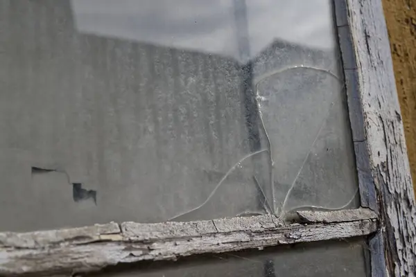 Kırık camlı ve soyulmuş boyalı yıpranmış bir pencerenin yakın çekimi. Toz ve kir camda görünüyor, bulanık bir arkaplanı yansıtıyor..