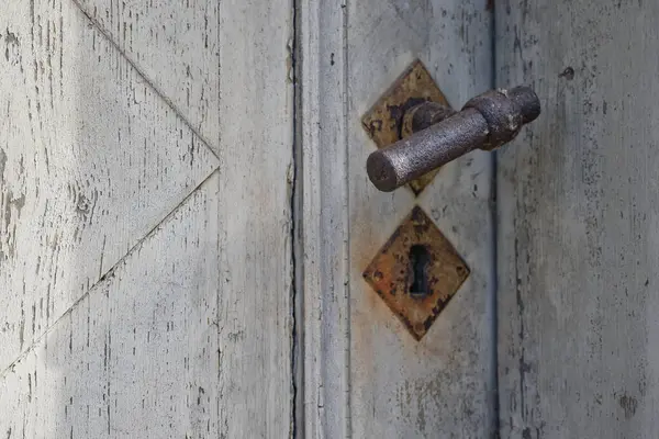 Eski ahşap bir kapıya yakın çekim, yıpranmış bir yüzeyi, paslı bir metal sapı ve elmas şeklinde bir anahtar deliği var..