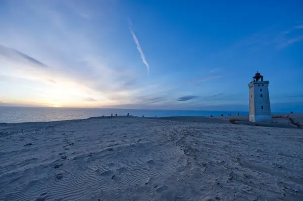 Gün batımında deniz feneri Lyngvig Fyr 'in sahnesi. Güneş ufukta batarken gökyüzü yumuşak mavi ve turuncu renklerle boyanır. Uzaktan insanların siluetleri görülebilir, sakin atmosferin tadını çıkarırlar..