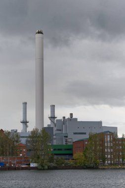 Bir su kütlesinin yanında uzun bacaları olan büyük bir endüstriyel enerji santrali. Gökyüzü kara bulutlarla kaplı ve Stadtwerke Flensburg yakınlarında ağaçlar ve binalar var..