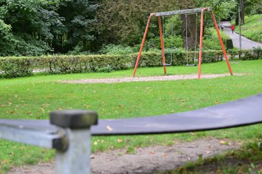 Flensburg Ostseebad 'daki sakin bir parkta yemyeşil çimlerle çevrili bir salıncak seti var. Arka planda bir patika görülebilir, yürüyen bir kişi ile.