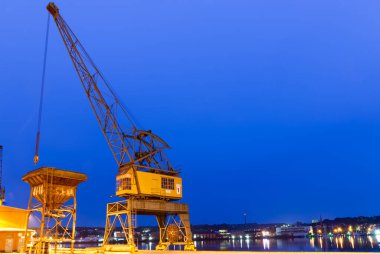 Alacakaranlıkta şehrin ışıklarıyla aydınlatılan büyük bir endüstriyel vinç rıhtımın yanında duruyor. Gökyüzü derin bir mavidir, suyun yüzeyini yansıtır, sakin bir atmosfer yaratır..